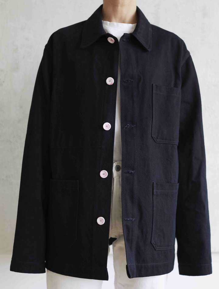 Model image of black workwear inspired jacket over an all-white outfit and white wall background